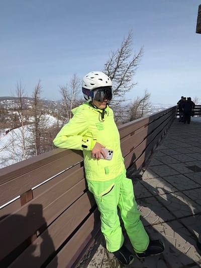 An adult female in full ski attire is depicted standing on a paved observation deck in Zelenaya Polyana, Russia, overlooking a snowy mountain landscape under a clear sky. The subject wears a white ski helmet with dark, mirrored goggles covering her eyes, a fluorescent yellow-green hooded ski jacket with black detailing and "COO ZOK" text, and matching ski pants with black accents and zippered pockets. Her lower legs are obscured by black ski boots or liners. She is leaning with her left arm resting on a dark brown wooden railing that extends along the deck. In her right hand, she holds a light-colored smartphone with a triple-camera system, positioned against her hip. In the far right background, several other individuals in winter clothing are standing on the same paved surface beyond the railing. The surrounding environment consists of snow-covered slopes, sparse deciduous trees with bare branches, and some evergreens. A marked ski run is visible in the mid-ground to the left. The ground surface is composed of grey rectangular pavers, and bright sunlight casts distinct shadows.