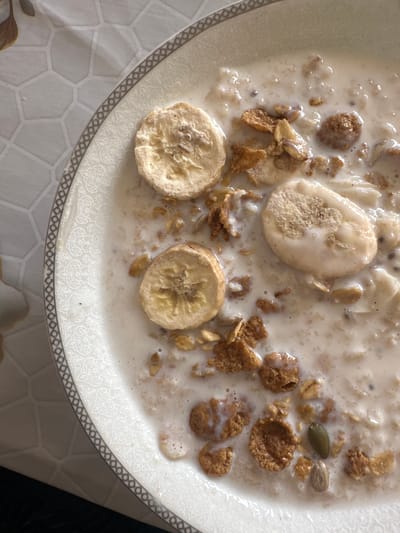 A close-up shot shows a bowl of cereal with sliced bananas in a milky liquid, likely milk or yogurt. The cereal appears to be a mix of flakes and granola. Two pumpkin seeds are also visible. The bowl rests on a tablecloth with a geometric pattern. The location of Agarak, Armenia is not evident from the visual data.