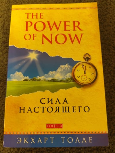 This is a close-up, eye-level, indoor photograph of the book cover for "The Power of Now" by Eckhart Tolle, with the title also displayed in Russian: "СИЛА НАСТОЯЩЕГО" (SILA NASTOYASHCHEGO). The cover is predominantly yellow with red lettering. The top portion of the cover features the English title "THE POWER OF NOW". Below this, a torn-edge landscape image depicts a bright sun with rays shining down on a green field with scattered trees and a blue sky filled with fluffy white clouds. To the right of this landscape, a vintage pocket watch with Roman numerals is superimposed. The Russian title is printed in large black letters below the landscape. A small red rectangle with white Russian text "СОФИЯ" (SOFIYA), which is the publisher's imprint, is located to the right of the Russian title. The bottom of the cover is a dark blue band with the author's name in white Cyrillic letters: "ЭКХАРТ ТОЛЛЕ" (EKKHART TOLLE). The book is lying on a dark, textured surface, likely carpet. There is no discernible location in the city of Schwäbisch Gmünd, Germany.