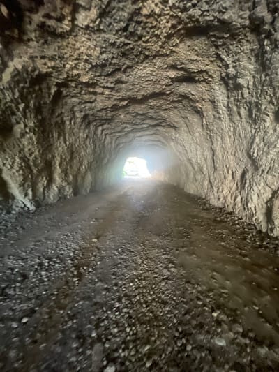 The image shows a narrow, rough-hewn tunnel or passage, likely underground or carved into rock. The walls are uneven and textured, with visible striations and a gritty appearance. The passage curves slightly, leading towards a bright light source at the end, suggesting an exit. The floor appears to be uneven and possibly damp, with small stones and dirt. The overall impression is one of an ancient or natural passage, possibly a cave, an old mine shaft, or an aqueduct. The lighting at the end of the tunnel is bright, hinting at daylight or a strong artificial light source outside. The scene conveys a sense of mystery and exploration, with the contrast between the dark, rough interior and the bright exterior. There is no text visible in the image.