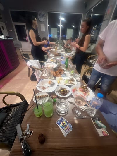 The image depicts an indoor scene, likely a restaurant or bar, with a long table laden with used plates, glasses, and bottles. Several individuals are present, some seated, others standing. The table is cluttered with remnants of a meal, including plates of food, cutlery, glasses containing drinks, and an ashtray. There are also a few personal items on the table, such as keys, a pack of cards, and a water bottle. The environment appears to be dimly lit, with artificial lighting and a view of the outdoors through a large window in the background. The presence of food and drinks suggests a social gathering or a meal in progress. The specific location in Zlataritsa, Bulgaria, is not visually discernible from the provided image.