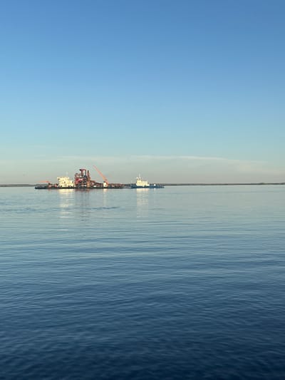 The image shows a body of water, possibly a wide river or lake, with a barge or construction platform in the distance. The sky is clear blue, suggesting a calm day. The water surface has slight ripples. The location is likely Soroch'i Gory, Russia.