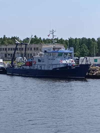 The image captures a vessel named "DMC SAILOR" docked in the harbor of Pāvilosta, Latvia. The boat, with a dark blue hull and a lighter blue superstructure, is positioned in the foreground, partially obscuring the water. The vessel is equipped with an A-frame structure at its stern and a smaller crane-like apparatus on its deck. Red lifebuoys are visible on the side of the superstructure, and the vessel's name is clearly displayed on its bow.

In the background, a low-rise, utilitarian building with multiple windows lines the shore. Behind the building, a dense line of green trees forms a natural backdrop. To the right of the vessel, several trailers are parked near a rocky embankment. The sky above is a clear, pale blue, suggesting fair weather. The water in the foreground exhibits a textured surface with numerous ripples, indicating a calm, yet active, harbor environment. No people are clearly visible in the image.
