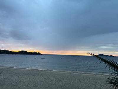 The image depicts a serene beach scene at dusk, likely in Lệ Uyên, Vietnam. A sandy beach occupies the foreground, transitioning into a calm, blue ocean that stretches towards the horizon. In the distance, silhouetted hills and islands are visible against a sky painted with the soft hues of sunset, transitioning from pale orange near the horizon to a muted blue and grey with scattered clouds. Several small boats are dotted across the water, suggesting a fishing or coastal community. The overall atmosphere is tranquil and peaceful, capturing the quiet beauty of the coastline as day gives way to night.