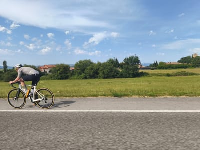 A cyclist is riding on a paved road with a white line marking the lane. The cyclist, wearing a helmet, grey t-shirt, and black shorts, is positioned on the left side of the frame, moving from left to right. The bicycle is white with black handlebars and wheels. To the right of the road is a grassy field with a line of trees and buildings in the background. The sky is blue with scattered white clouds. The location appears to be rural, possibly in Spain given the architectural style of the buildings.