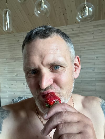 A man is shown in a close-up, self-portrait style photograph, holding a strawberry to his mouth. He has graying hair, blue eyes, and is shirtless, wearing a gold chain. The background appears to be an indoor setting with a wooden paneled ceiling and a wall made of light-colored, horizontal planks. The environment suggests a residential setting, possibly a room with a rustic or modern aesthetic, with pendant lights visible on the ceiling. The image does not provide any specific indicators of the city of Aleksandrovka, Russia.