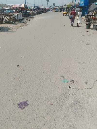 The image presents an eye-level view of a wide, unpaved or gravely grey road under an overcast sky, located in a market area of Mgbuoshimini, Nigeria. The foreground of the road exhibits scattered debris including small colored fragments (e.g., purple, green) and dark smudges. Along the left side of the road, a continuous line of market stalls with rough wooden frames and diverse tarpaulin or umbrella coverings extends into the distance, with litter visible on the ground beside them. A dark-colored sedan is parked among these stalls further back. On the right side of the midground, two adult females are walking away from the camera: one wears a dark red top and a patterned skirt with a dark shoulder bag, and the other wears a light-colored dress. Beyond them on the right, a blue building with partial white lettering is visible, adjacent to a market stall displaying produce. Further down the road, a yellow tricycle (Keke Napep), a light-colored car, and a yellow wheelbarrow are present. Utility poles with overhead wires line the street, and a water tower is discernible in the far background.