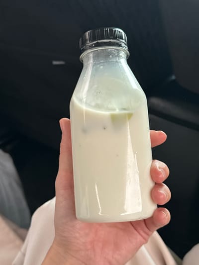 A hand holds a bottle of white liquid, possibly yogurt or milk, containing small green and black particles. The bottle has a black cap. The background shows a dark, possibly car interior, and a glimpse of a white garment. The location, Meshcheryaki, Russia, is not evident from the image.