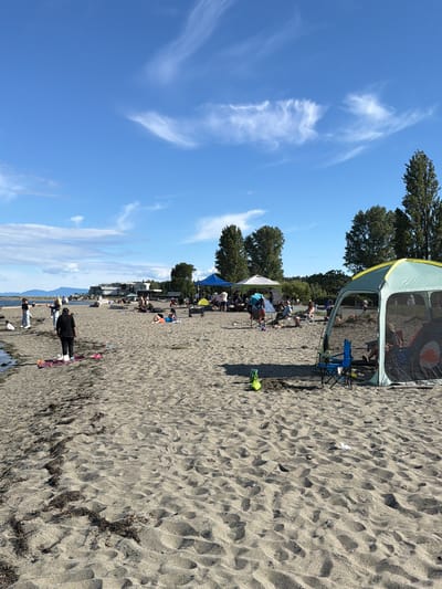 This image depicts a sunny day at a beach in the city of Delta, Canada, likely Crescent Beach, given the presence of a sandy shore, calm water, distant buildings, and a line of trees. A variety of people are engaged in beach activities. In the foreground, a person is standing on a pink towel on the sand, facing away from the camera. To the right, a large, open-sided tent with a yellow trim provides shade for several individuals, including one person in a hat visible inside. Two blue camping chairs are set up outside the tent. Further back, several other tents and canopies are visible, with groups of people relaxing, sitting, and conversing. Some individuals are lying on blankets or chairs on the sand, while others are standing and interacting. The beach is covered in sand with scattered seaweed and driftwood. The sky is a clear blue with a few wispy white clouds. In the distance, there are buildings and a line of trees, with mountains faintly visible on the horizon.