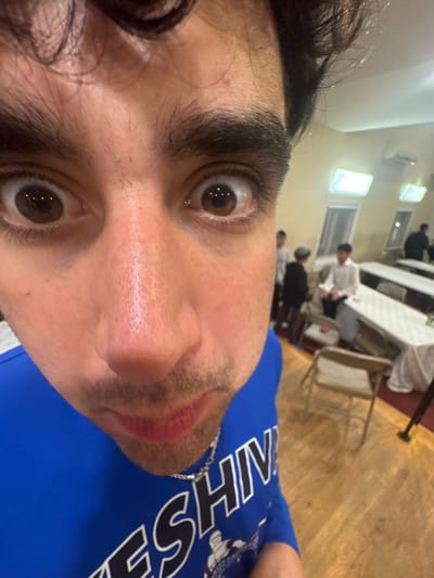 A close-up, wide-angle image depicts a young male with dark hair, prominent eyebrows, and wide brown eyes looking directly into the camera. His face appears flushed, and he wears a blue long-sleeved shirt partially displaying the white letters "YESHIVA," along with a silver chain necklace. The background reveals an indoor communal hall, likely in Bergenfield, United States, given the context. The hall features light-colored walls, multiple rectangular fluorescent light fixtures, an air conditioning unit, and a wooden floor. Several long tables covered with white tablecloths and beige folding chairs are arranged in the space. Multiple young males are visible in the background, some seated at the tables, one wearing a dark cap, indicating a gathering within a Jewish educational or communal institution.