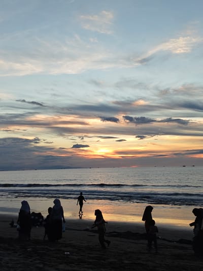 The image depicts a beach scene at dusk in Uleekareung, Indonesia. Silhouetted figures of people, including adults and children, are scattered across the sandy shore. Some individuals are walking, while others appear to be gathered. The ocean is visible with gentle waves lapping at the shore. The sky is filled with dramatic clouds, painted with hues of orange, pink, and blue, indicating a sunset. A few small boats are visible in the distance on the water. The overall atmosphere is serene and twilight.