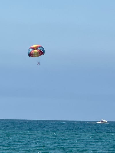 The image depicts a bright, clear day over the sea. In the sky, a colorful parasail, with red, yellow, and blue sections, is visible, carrying two people suspended beneath it. Below, on the turquoise water, a white speedboat is traveling, leaving a white wake behind it. The horizon is visible, separating the sea from the pale blue sky. The scene is likely Vityazevo, Russia, a resort town known for its beach activities.