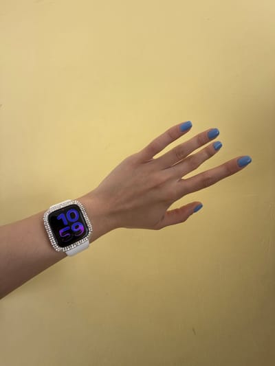 A left human hand, with fair skin and fingers slightly splayed, is depicted against a plain, solid yellow-ochre wall in Molodezhnyi, Russia. The fingernails are painted with light blue polish. A smartwatch, identified as an Apple Watch, is worn on the wrist with a white silicone strap. The watch face is enclosed within a clear protective case adorned with two rows of small, clear rhinestones. The watch screen displays the time "10 59" in dark blue digital characters. Minor scuff marks are visible on the yellow wall.