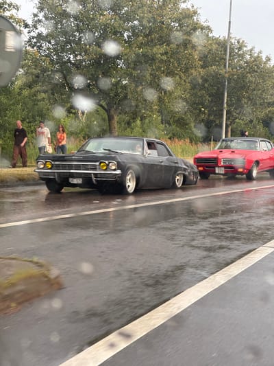 In Falkenberg, Sweden, on a wet road, two vintage American cars are parked. The car in the foreground is a black, four-door sedan with yellow headlights. It has a license plate that reads "CHF 075". Behind it, to the right, is a red two-door coupe with the license plate "DSC 277". The red car appears to be a Pontiac GTO. Three people are standing on the sidewalk to the left of the black car. One is a man in a dark shirt and brown pants, another is a man in a white t-shirt and jeans, and the third is a woman in an orange and white top and jeans. The trees in the background are full of green leaves, and the sky is overcast, suggesting it might be raining or has recently rained. The road surface is wet and reflective, indicating damp conditions.