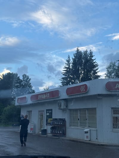 A man holding a child walks away from the camera on a wet asphalt surface in front of a white, corrugated metal building. The building has a red sign with white lettering that reads "ЛУКОЙЛ" (Lukoil). There are several red oval signs on the building, some with white arrows. Windows with metal grates are visible on the building. To the right of the entrance, there is a rack of oil cans. In the background, there are several tall pine trees and a cloudy blue sky. The scene appears to be a gas station or convenience store in Chernovskoye, Russia.