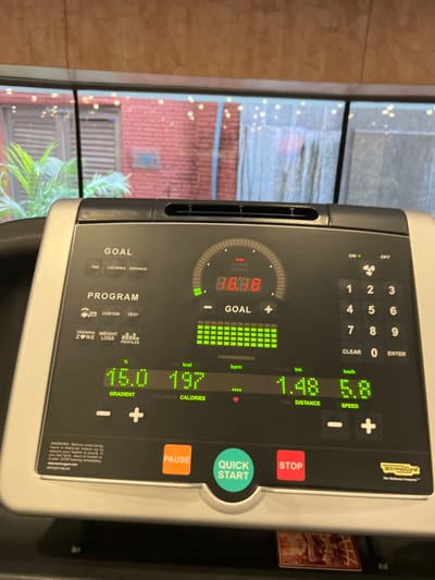 The image presents a close-up, eye-level view of an active Technogym treadmill console, positioned indoors in Noida, India. The digital display is illuminated, showing various workout metrics. A central red LED clock displays "08:08", likely representing elapsed time. Below this, a green bar graph indicates progress towards a "GOAL". Further metrics include a "GRADIENT" of 16.0%, "CALORIES" at 197 kcal, "DISTANCE" at 1.48 km, and "SPEED" at 5.8 km/h. A small red heart icon is visible under "BPM". The console features orange "PAUSE", green "QUICK START", and red "STOP" buttons, along with a numeric keypad and branding for "Technogym The Wellness Company".
Beyond the treadmill console, a large window reveals an outdoor environment. This includes a building with reddish-brown brickwork, partially obscured on the left by green foliage, resembling a palm plant. Numerous small, bright, scattered lights are visible across the background, suggesting decorative string lights. A grey, vertically hanging fabric or curtain is also partially discernible on the right side of the outdoor view.