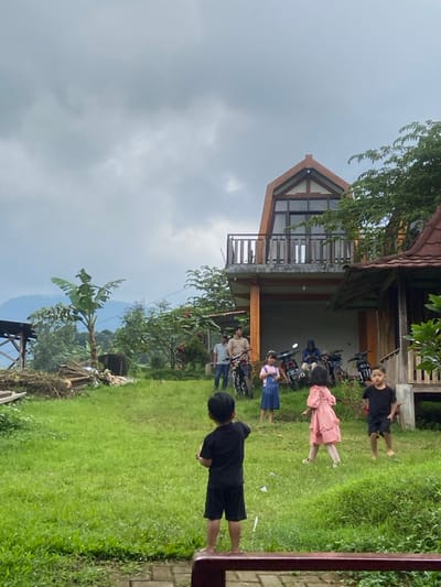 The image depicts an outdoor scene in Penanggungan, Indonesia, under a heavily overcast sky. In the foreground, a dark red railing partially obscures the bottom edge. Four children are present on a sloping green grassy area. A young boy in a black t-shirt and shorts stands with his back to the viewer, facing towards the center. To his right, a girl in a pink ruffled dress is walking to the left. Further right, another young boy in a black t-shirt and shorts moves towards the left. A girl in blue denim overalls stands stationary to the left of the girl in pink.
In the midground, towards the right, stands a multi-story building featuring an A-frame upper section with large windows and a balcony with wooden railings. The lower level is open, revealing a concrete area where several motorcycles are parked. Three adult males are visible near the motorcycles; one in a light beige shirt and another in a light blue shirt stand upright, while a third is seated on a motorcycle. To the immediate right of the main building, a smaller structure with a reddish-brown tiled roof is partially visible.
The left side of the scene includes a tall banana tree, a pile of wood debris, and a wooden frame structure. Distant mountains or hills are visible through a haze in the background beneath the cloudy sky. The ground throughout the visible area is primarily green grass, indicating an uneven, natural terrain.