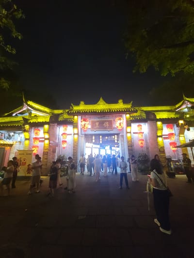 The image depicts a nighttime scene at the entrance to a traditional Chinese-style building, likely a market or entertainment district, in Wuhou District, China. The structure features ornate, yellow-lit eaves and is adorned with numerous red lanterns, some of which are lit. A sign with Chinese characters is visible above the entrance archway. A group of people, dressed in casual attire, are gathered in front of the building, some walking and others standing. The ground is paved, and trees with green foliage are visible in the upper corners of the frame, silhouetted against the dark night sky. The lighting is artificial, casting a warm glow on the building and the surrounding area, with some areas of shadow.