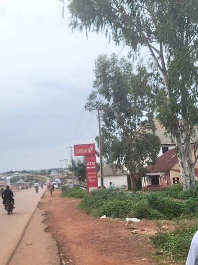 The image depicts a roadside scene in Bokkos, Nigeria, likely on a cloudy day. A paved road, with some visible cracks and disrepair, curves to the left and recedes into the distance. Several motorcycles are present on the road; one in the foreground is ridden by a person wearing a dark shirt, while others are further down the road. A person in a light blue shirt is visible standing on the side of the road.
To the right of the road, there is a red sign with "UBA" and "ATM" written on it, indicating a bank. Adjacent to the sign are several buildings with red and white corrugated iron roofs, interspersed with greenery. Large trees with sparse foliage line the area between the road and the buildings. Some debris, including rocks and white plastic bags, are scattered on the unpaved area to the right of the road. The background shows a slightly hazy view of more buildings and hilly terrain.