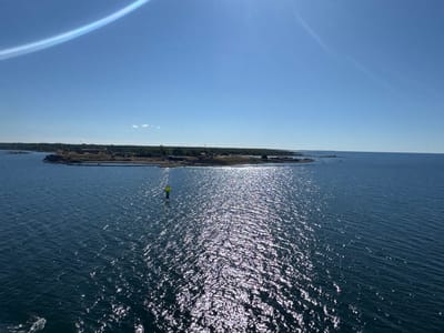 The image shows a view of the sea towards the island of Drottningskär, Sweden. The sky is clear blue with a bright sun creating a lens flare effect in the upper left corner. A long, thin cloud or atmospheric phenomenon stretches across the sky. The sea is dark blue with the sun's reflection creating a shimmering, sparkly surface. In the distance, the island of Drottningskär is visible, characterized by its rocky terrain and a cluster of buildings, possibly part of a fortress or historical site. A green and yellow buoy is present in the water. The overall scene depicts a calm, sunny day at sea with a view of the Swedish coastline.