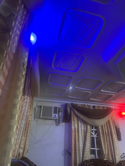 The image depicts an interior scene, likely a living room, with a focus on the ceiling and a window area. The ceiling features a decorative coffered design, illuminated by blue LED lights. A small, round blue light fixture is visible near the top left of the frame, casting a bright blue glow. To the left of the window, a window air conditioning unit is mounted on the wall. The window itself is draped with ornate, gold-colored curtains, featuring a patterned overlay and layered fabric accents. A dark brown and light beige draped valance hangs above the curtains. The wall appears to be painted a light color. To the right of the window, a red light is visible, suggesting additional mood lighting. The overall impression is of a room decorated with attention to lighting and soft furnishings. The location in Kporva, Nigeria is not evident from the visual information in the image.