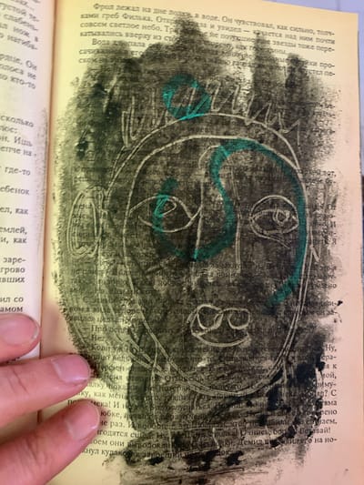 This is a close-up, eye-level, outdoor photograph of a page from a book that has been defaced with drawings. The book page is yellowed and textured. The text is in Russian and is partially obscured by the drawings and a human hand. The drawings are abstract and appear to be a stylized face, rendered in black and green crayon. The face has large, circular eyes with lines inside, a large nose, and a mouth with circles. The head is outlined with a green crayon, and there is a green zigzag line through the center of the face. A human hand with short, clean fingernails is visible in the bottom left corner of the image, holding the book open. The background of the drawing is textured and appears to be made with a rough, smudged black material, possibly charcoal or a dark crayon. The image does not contain any visual elements that would allow for the determination of a location in Ganino, Russia.