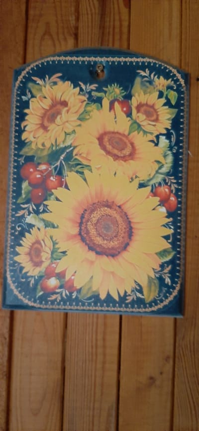 This is a close-up, top-down shot of a rectangular decorative tray with rounded corners, featuring a painting of sunflowers and cherries. The tray has a dark blue background and is bordered with a decorative pattern of small pink dots and light beige swirls.
The main subjects of the painting are several large sunflowers in various stages of bloom, rendered in vibrant yellows and oranges with dark brown centers. Interspersed among the sunflowers are clusters of bright red cherries. Green leaves and stems complete the floral and fruit motif.
The tray itself is positioned on a wooden surface, likely a table or shelf, composed of horizontal wooden planks with a warm, light brown hue. The lighting appears to be natural, possibly from a window, casting a soft glow on the scene. The image conveys a rustic, homey aesthetic. There is no visible text in the image. The context provided, "Былина Снт, Russia," suggests the location where this tray might be found. No people are visible, and no specific event or activity is depicted, other than the decorative nature of the object.