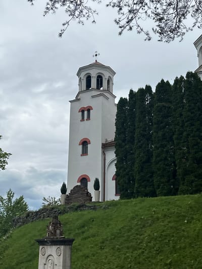 The image shows a white church tower with a red-brick trim, standing against a cloudy sky. To the right of the tower is a tall, dark green tree, and further to the right is a grassy area with a stone wall and a tombstone. A bare tree branch is visible in the upper left corner. The church is located in Barziya, Bulgaria.