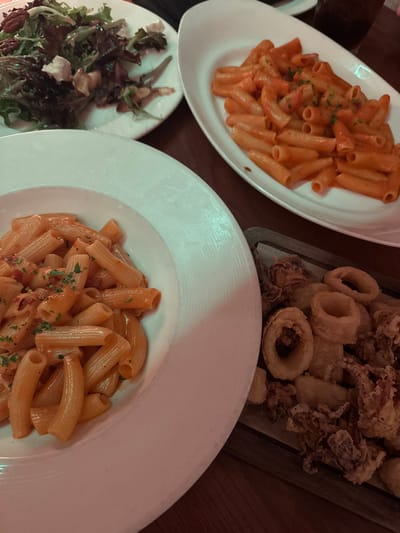 A table setting features three dishes. Two white plates hold penne pasta with a creamy tomato sauce, garnished with parsley. A smaller plate displays a mixed green salad with feta cheese and pecans. Fried calamari is arranged on a wooden board, partially visible. The composition suggests a shared meal or appetizer selection.