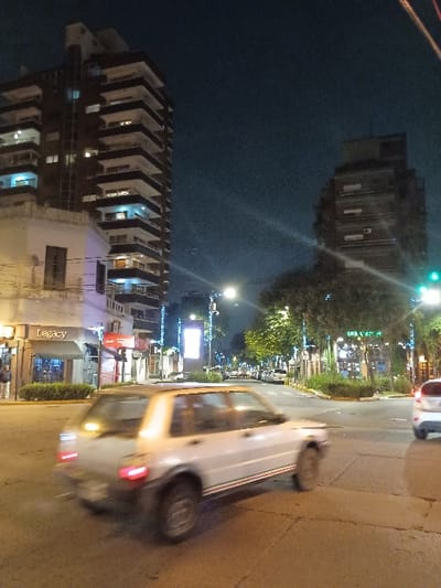 The image shows a street intersection in Victoria, Argentina, at night.  The intersection is located at the coordinates -34.453, -58.539.  There are two buildings on the left side of the image: a tall building with a sign that says "Legacy" on the ground level and a shorter building with a sign that says "Mexico" on the ground level.  There is a white car driving through the intersection.  The car is being driven by a person that is not visible. The surrounding area is dark, but streetlights illuminate the street and the cars driving in both directions.  The buildings and streetlights appear to have some Christmas decorations, though this is unclear.  The car's speed is not clear, but it appears to be moving at a slow speed.  The street surface is asphalt.  The sky is clear and dark, and the buildings are visible in the background.  There are no other people visible in the scene.  The image was likely taken by someone standing on the sidewalk next to the "Legacy" building.  The image's perspective is from slightly above the street level, making it look like the car is driving toward the viewer.  There is a streetlight shining on the buildings in the background, and there are several other streetlights illuminating the street.  The streetlights are bright and cast long shadows.