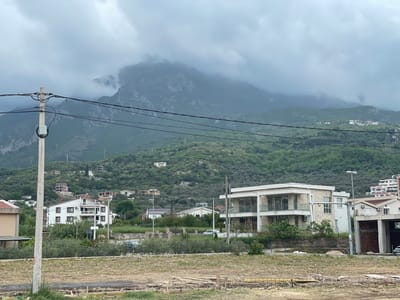 The image presents a sweeping view of a residential area in a mountainous region, likely Tomba, Montenegro, under an overcast sky.
In the foreground, there is a flat, uncultivated plot of land, appearing dry and dusty, with visible wooden planks and rebar, indicating either preparing for or being a site of construction. A prominent concrete utility pole with a dense network of black power lines extends from the left side of the frame, traversing the mid-ground.
The middle ground showcases a cluster of residential buildings integrated into a verdant, hilly landscape. The houses display varying architectural styles, including a contemporary multi-story building on the right with large windows and balconies, and a more traditional white house with brown window frames on the left. Lush green trees and dense bushes are abundant, covering the hillsides and surrounding the residences. Several smaller houses are scattered further up the slopes. A silver car is partially visible near a road in the mid-right, and another dark vehicle is barely seen on the far right.
The background is dominated by imposing, rugged mountains. Their lower and mid-sections are thickly covered with green vegetation, while their peaks and upper slopes are shrouded in heavy, gray clouds or mist, creating a dramatic and somewhat ethereal backdrop.
The time of day appears to be daytime, but the weather is characterized by a completely overcast sky with thick, gray clouds, suggesting a muted, diffused light. The scene depicts a quiet moment in a naturally scenic area undergoing development, with no people visible and no legible text.