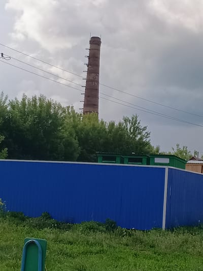 The image captures a view of a tall, weathered brick smokestack rising against a cloudy sky in Tolbazy, Russia. The smokestack features visible rungs ascending its exterior and appears to have the number "19" painted on it. In the foreground, a vibrant blue corrugated metal fence spans horizontally across the frame, separating the viewer from the background. Behind the fence, lush green trees and foliage form a dense line, partially obscuring structures including a low-lying green building with rectangular windows and a segment of a wooden structure to the right. The ground in the immediate foreground is covered in green grass, with a green pole-like object visible on the left. Power lines are strung across the upper portion of the image.