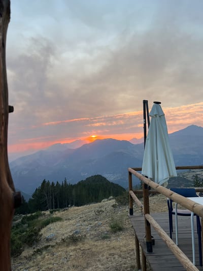 The image depicts a mountainous landscape at sunset, viewed from a wooden deck. The sun is partially visible behind a range of mountains, casting a warm orange glow across the sky and illuminating the clouds with shades of pink and orange. In the foreground, a white patio umbrella is folded and standing upright, attached to a black pole. A wooden railing borders the deck, and a blue chair and a white table are visible on the deck. The mountains in the background are layered, with the closest ones covered in dark green pine trees, and further mountains fading into a hazy blue. The ground on the deck and the slope leading away from it is covered in dry, golden grass and scattered rocks. The scene suggests an outdoor seating area in a high-altitude location, likely a restaurant or viewpoint overlooking the valley. The village of San Juan de Plan is visible in the distance in the valley below the mountains.
