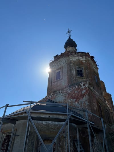 The image shows a low-angle view of a partially ruined brick building in Шуран, Russia, set against a clear blue sky. The building's facade is weathered, with significant deterioration of the brickwork and plaster. Scaffolding supports sections of the building. The sun shines from behind the structure. A dome with a cross sits atop a square tower.
