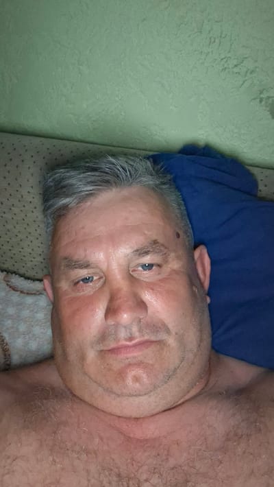 A selfie is taken from a low angle by a fair-skinned adult male with graying hair, who is lying down. He is shirtless, showing a hairy chest, and resting his head on a blue pillow. The background consists of a textured, light green wall and a portion of a patterned cushion. The man's face is the primary focus, with his blue eyes looking directly at the camera. He has some facial stubble and a mole on his left temple. The overall lighting is dim, suggesting an indoor setting, possibly a bedroom. There is no visible indication of the location being in the city of Voinova, Moldova.