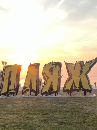 A large, yellow and black sign with gothic-style lettering reads "ПЛЯЖ" (which translates to "BEACH" in Russian). The sign is positioned on a sandy area with a grassy foreground, suggesting a beachside location. In the background, a body of water is visible, reflecting the warm hues of the sunset. The sky is filled with soft, diffused light, with some wispy clouds and a contrail from an aircraft. Several people are scattered in the background, appearing to be enjoying the sunset or an event. One person is standing near the sign, while others are more distant. The overall scene conveys a relaxed, end-of-day atmosphere. The location is identified as Surakovo, Russia.