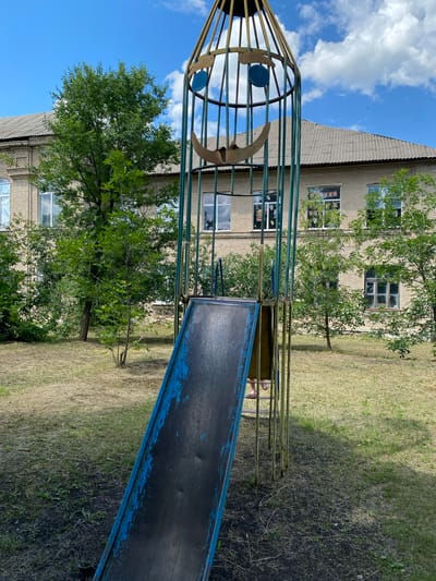 A child's legs and feet, wearing pink sandals, are visible near the base of a tall, narrow playground structure. The structure is made of metal poles and has a slide attached. The slide is blue and worn, with some paint peeling off. In the background, a light-colored brick building with multiple windows stands behind trees. The ground is covered with dry grass and patches of dirt. The sky is blue with scattered white clouds. The scene appears to be an outdoor playground area, likely in or around Markivka, Ukraine.