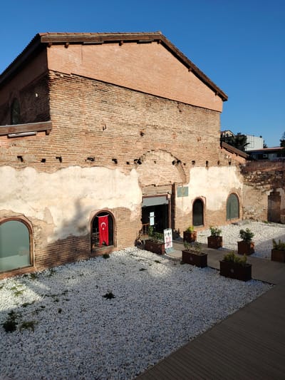 An outdoor, sunlit view captures a historic brick building with a courtyard in Iznik, Turkey. The main subject is a sturdy, reddish-brown brick structure, partially covered with peeling light-colored plaster, revealing the underlying brickwork. The building features several arched window openings, some glazed, and a central arched entrance. A vibrant red Turkish flag, adorned with a white star and crescent, hangs prominently in one of the arched window openings on the left side of the facade.

The building's roof has classic terracotta tiles and visible wooden eaves. In front of the structure is a courtyard paved with numerous small, white pebbles. Interspersed within the pebble area are dark wooden planter boxes containing small green plants. A modern-looking wooden decking path runs along the right side and front of the courtyard.

The sky above is a clear, brilliant blue, indicating fair weather. Strong shadows cast by the building suggest it is either mid-morning or late afternoon. To the left, an oval-shaped window is visible on the side of the building. Above the main structure, in the distance, parts of other buildings and some greenery, possibly palm trees, can be seen.

Near the main entrance, a white sign with several pictograms (suggesting health or safety guidelines like social distancing and mask use) is visible, though specific text is unreadable. A small blue plaque is also affixed to the wall above the entrance, but its text is likewise indiscernible. There are no people visible in the image, creating a serene and timeless atmosphere.