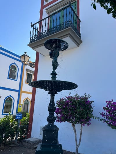 A black wrought iron fountain with a cherub statue sits in a paved courtyard in front of a white building with a green-trimmed balcony. The fountain is spraying water and there is a small, flowering bush on either side of the fountain. To the left of the fountain is a building with blue and white trim and a sign that reads "peatonal area." The coordinates provided (27.818, -15.764) correspond to Lomo Quiebre, Spain.