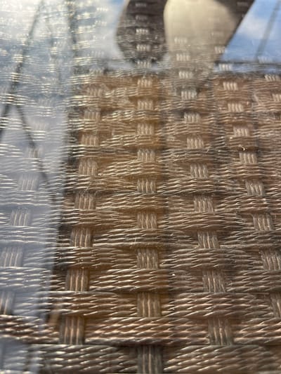 The image is a close-up, abstract view of a woven textured surface, likely an outdoor furniture material such as rattan or wicker. The texture is characterized by a tight weave of individual strands, creating a pattern of interlocking rectangles and diagonal lines. The lighting suggests it is daytime, with sunlight casting strong highlights and shadows across the woven material, emphasizing its three-dimensional texture.
In the upper left quadrant, a blurred reflection of what appears to be an outdoor scene is visible. This reflection shows a light-colored object, possibly part of a chair or table, against a bright, possibly blue sky. This suggests the object with the woven texture is outdoors, likely in a setting such as a patio, balcony, or garden. The reflection is distorted by the texture of the material it's cast upon, creating a shimmering, almost watery effect in some areas.
The overall impression is one of detail and texture. The focus is entirely on the materiality of the woven surface and the play of light upon it. There are no people visible, nor any specific discernible event taking place beyond the passive presence of the object in its environment. The image is devoid of any visible text. The context provided, "Azov, Russia," is not visually evident in the image itself but is understood as the location of the object depicted.