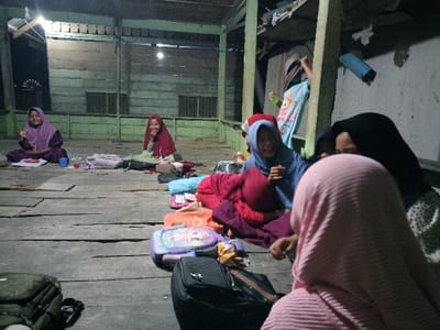 The image captures an indoor scene within a wooden building in Cutneuheun, Indonesia, illuminated by artificial overhead lighting. The floor is composed of worn wooden planks, and the walls are constructed from light green/blue wooden boards with visible support beams. A group of eight individuals, primarily young females wearing hijabs, are present.
In the foreground, a person in a pink ribbed hijab is seen from behind, facing right. Adjacent, a person in a black hijab and patterned top interacts with a young girl in a blue hijab and red jacket, who is smiling and gesturing with her right hand. A black backpack and a light purple lunchbox are on the floor near them.
In the mid-ground, two young girls are seated cross-legged on the floor. The girl on the left, in a purple hijab and dress, smiles at the camera and displays a two-finger gesture; a food container and a blue cup are in front of her. The girl on the right, in a red hijab and light top, also smiles at the camera, with a red cup and container nearby. Additional bags and personal items are scattered around them.
In the background, a person in a light blue top is partially visible behind a wooden pillar. The scene depicts an informal gathering or social interaction.