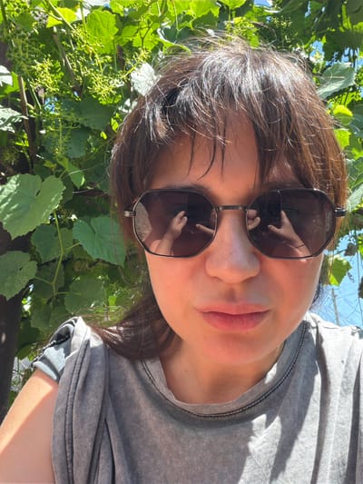 A woman is taking a selfie outdoors, with a blurred green vine and blue sky in the background. She is wearing sunglasses and a grey t-shirt. Her left arm and shoulder are visible, with sunlight casting shadows on them. The reflection in her sunglasses shows her hands holding the phone. The image is a close-up shot, focusing on her face and upper body.