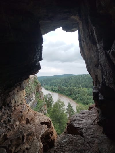 The image captures a view from within a cave, looking out onto a wide, winding river that flows through a densely forested valley. The cave entrance frames the landscape, with dark, textured rock formations on either side. The river, a muted brown color, reflects the overcast sky. The surrounding hills are covered in a thick canopy of green trees, with a few lighter-colored deciduous trees interspersed among the darker evergreens. The sky is cloudy, with a soft, diffused light illuminating the scene. There are no people or animals visible. The location is identified as a cave in the city of Kochkari, Russia.