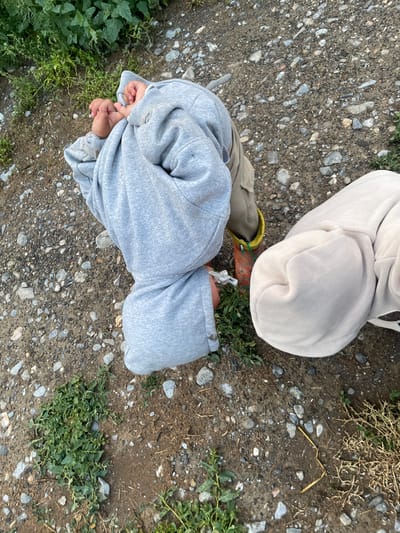 The image shows two children, one wearing a grey hooded sweatshirt and the other a beige hooded sweatshirt, crouching on a dirt and gravel path. The child in the grey sweatshirt is bent over with their head down, and their hands are clasped in front of them. They are wearing tan pants and orange boots with yellow accents. The child in the beige sweatshirt is also hunched over, with their head covered by the hood. The ground is covered in small stones, dirt, and patches of green vegetation. The scene appears to be outdoors in a rural or semi-rural setting, possibly in the city of Kara-Oi, Kyrgyzstan, given the context.