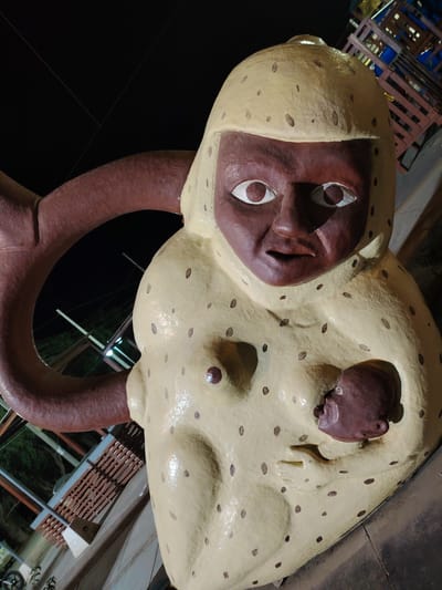 The image shows a statue of a woman with a baby located in the city of Trujillo, Peru. The woman has a brown face and large white eyes, and her body is light yellow with brown spots. She is holding a brown baby in her arms. Behind her is a brown circular structure. The background shows a dark night scene with some visible structures.
