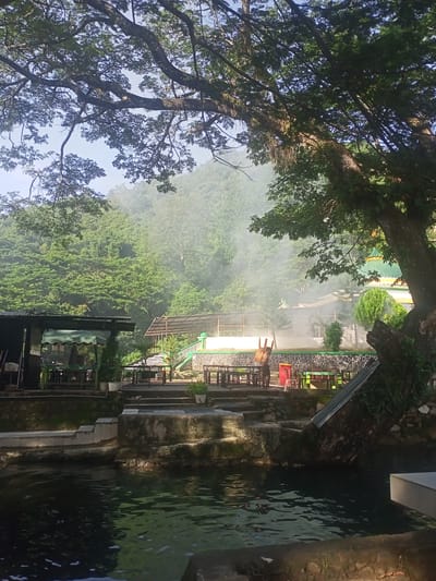 In the city of Geundring, Indonesia, a serene natural landscape unfolds, featuring a body of water in the foreground and lush green hills in the background. A large tree with extensive branches and foliage dominates the right side of the image, casting shadows and adding to the natural ambiance.
The scene appears to be a public area with several structures and amenities. To the left, a covered seating area with tables and chairs suggests a place for relaxation or dining. Adjacent to it, another covered structure with tables and chairs, possibly a restaurant or a resting spot, is visible. In the middle ground, a multi-tiered area with stone steps and platforms leads up to more seating arrangements.
A striking element is a building with a green dome and yellow accents, resembling a mosque or a similar religious structure, situated on the right side of the frame. In front of this building, there are manicured bushes and trees, indicating a well-maintained area.
A person is visible in the middle ground, carrying a chair and walking towards the right. The presence of mist or steam rising from the ground adds a mystical quality to the scene, particularly around the central structures. The water in the foreground is dark and reflective, with ripples on its surface, indicating movement. The overall atmosphere is tranquil and inviting, suggesting a popular spot for both locals and tourists.