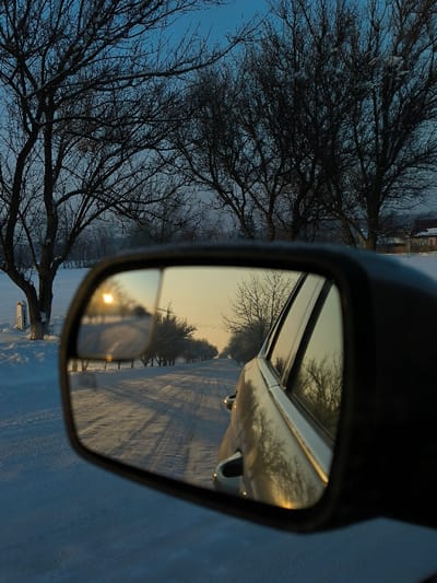 The image presents a serene winter landscape viewed from the interior of a vehicle, predominantly through its side mirror. The main subject is the car's right-hand side mirror, which reflects a snow-covered, straight road stretching into the distance. In the mirror's reflection, the sun, positioned low on the horizon, emits a bright, golden glow, casting long shadows across the pristine white snow. A line of bare, dormant trees flanks both sides of the reflected road, their branches appearing delicate against the warm light. The side of the car itself is also visible in the mirror, reflecting the same warm, winter sky and trees.

Outside the mirror's frame, directly in front and to the left of the car, are numerous large, bare trees, their intricate branches reaching towards a soft, blue sky. These trees appear dark and silhouetted against the lighter sky, with hints of snow or frost clinging to their branches. The ground outside the car is entirely covered in a thick blanket of snow. To the far left, a white post or marker stands in the snow near the road, with some faint, unreadable text or markings on its surface.

The activity suggests a quiet journey or stop along a snowy, rural road during either sunrise or sunset, indicated by the low angle and warm color of the sun. The setting is a cold, wintry environment in Khrinivka, Ukraine, characterized by snow and dormant vegetation. The mood evoked is one of peaceful solitude and the quiet beauty of winter.