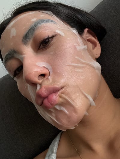 A woman with dark hair lies on a dark gray textured surface, possibly a couch, in a close-up, overhead shot. She is wearing a white sheet mask that covers most of her face, with cutouts for her eyes and lips. Her eyebrows appear darkened and shaped above the mask. Her lips are slightly parted and pouty, with a visible sheen from the mask's product. The mask has folds and wrinkles, suggesting it's conforming to the contours of her face. The background is mostly dark and out of focus, with a hint of a light-colored wall or surface visible in the upper left. The image does not provide any information that would suggest a location in the city of Imatra, Finland.