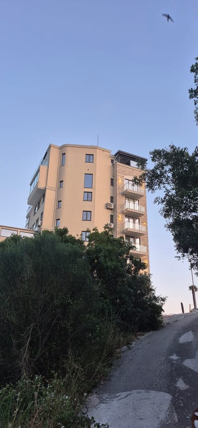 A modern, multi-story building with beige and stone facades stands against a clear, pale blue sky. The building features multiple balconies with glass railings and large windows. In the foreground, a winding asphalt road leads uphill, bordered by lush green vegetation. A bird is captured in flight in the upper right quadrant of the sky. The scene appears to be in Ivanovići, Montenegro, possibly during the late afternoon or early evening given the lighting.