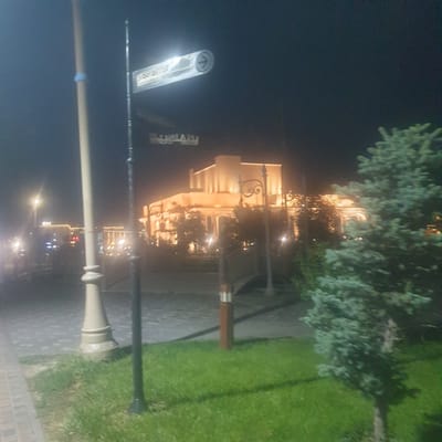 The image shows a nighttime scene in what appears to be Turkistan, Kazakhstan, indicated by the Cyrillic signage. A lit building with ornate architectural details stands in the background, partially obscured by trees. The scene is illuminated by streetlights, including one prominently in the foreground with a classical design. A sign for a metro station is visible. The environment includes a paved walkway, grass, and trees. There are no people visible in the image. The perspective is slightly tilted.